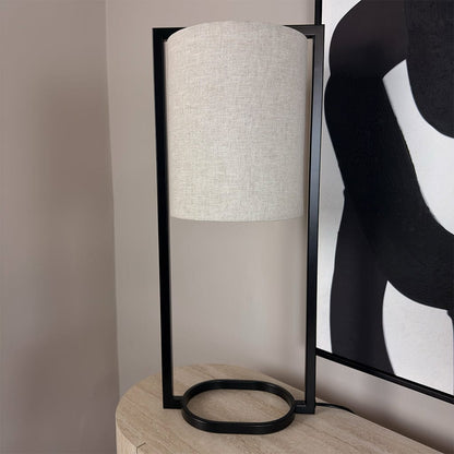 Table lamp with white shade and black base on a wooden surface, with abstract art in the background.