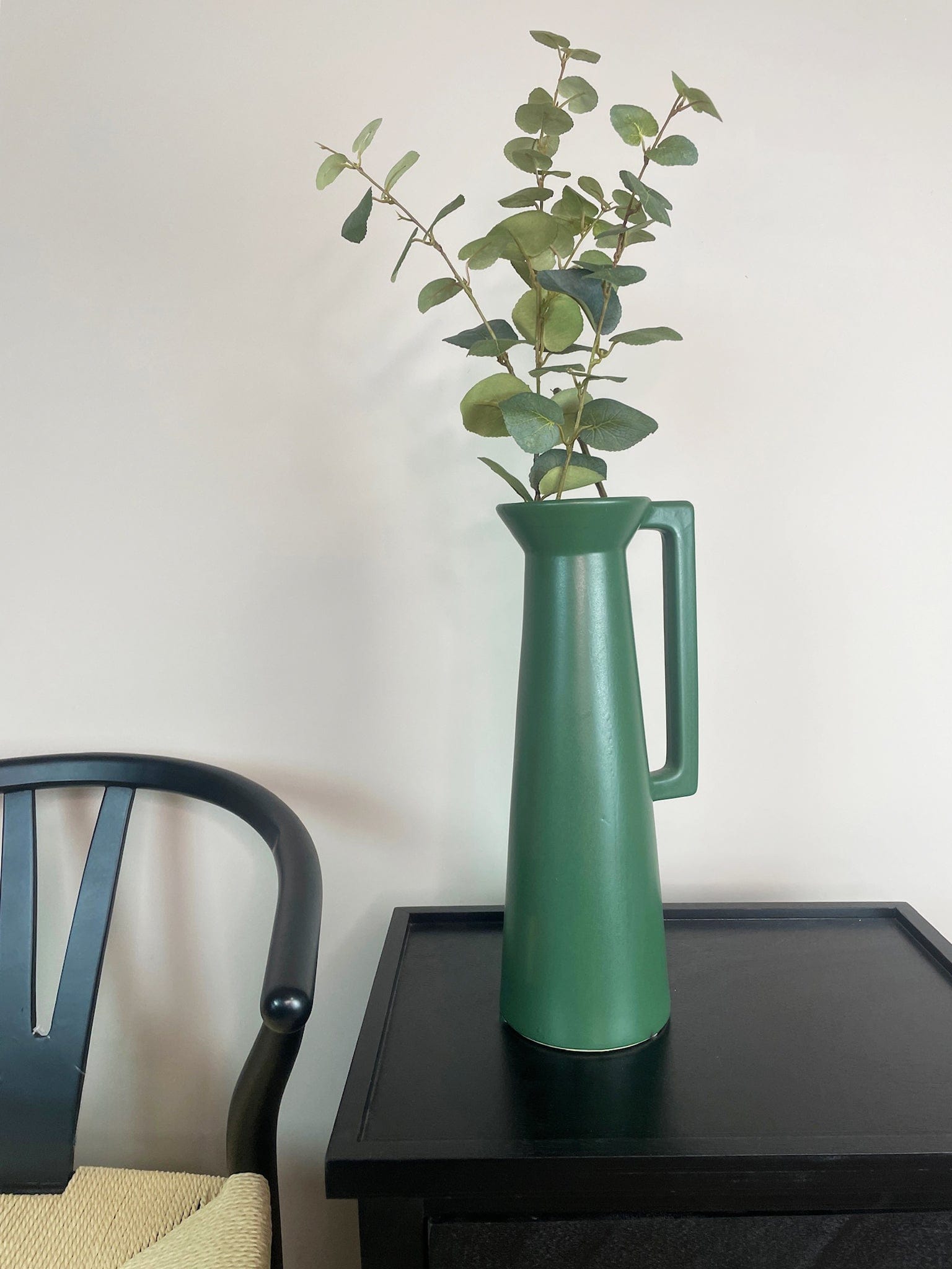 Green vase with a plant on a black table against a white wall