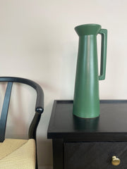 Green pitcher on a black surface next to a chair with a white wall background