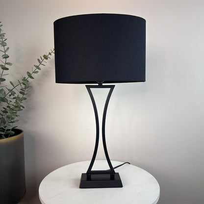 Black table lamp with a curved base on a white surface, with a plant in the background.