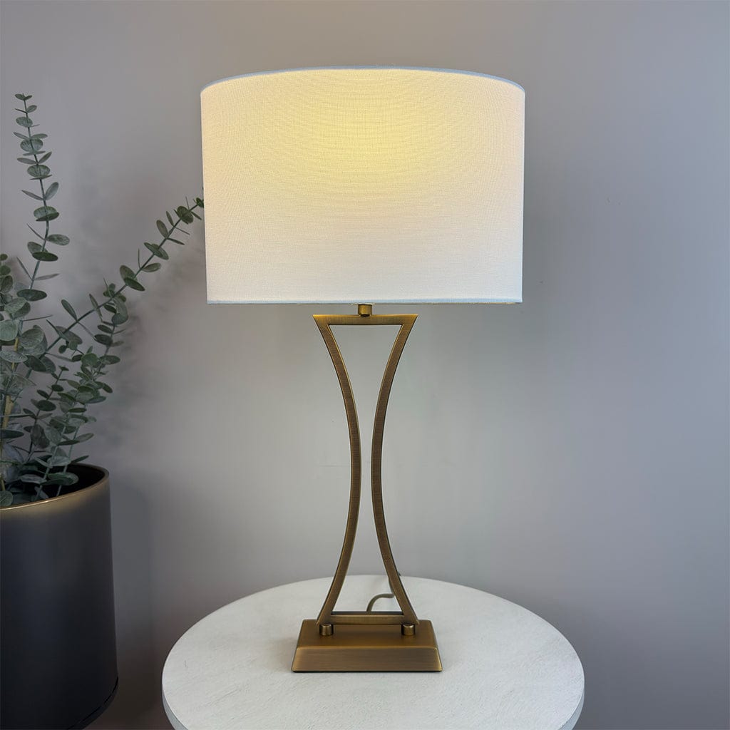 Table lamp with a white shade and gold base on a round white table, next to a plant against a gray wall.