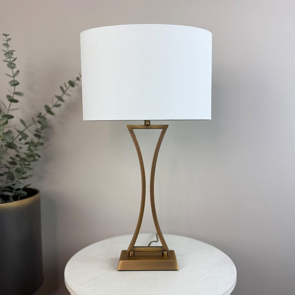 Table lamp with white shade and gold base on a round white surface, with a plant in the background.