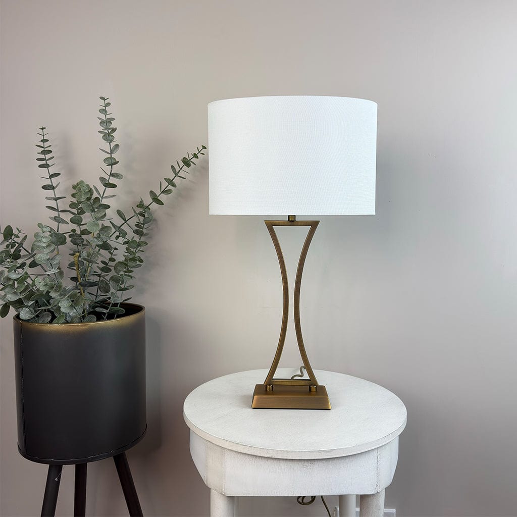 Table lamp with white shade on a round white table next to a potted plant against a plain wall.