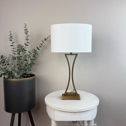 Table lamp with white shade on a round white table next to a potted plant against a plain wall.