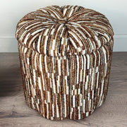 A round, tufted pouf ottoman with a textured vertical stripe pattern in brown, beige, and white, set on a gray wood floor against a white wall.