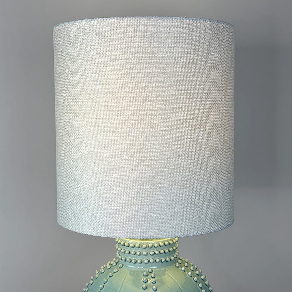A table lamp with a large cylindrical white textured fabric shade and a glossy pale green ceramic base decorated with raised bead details, shown against a plain gray background.