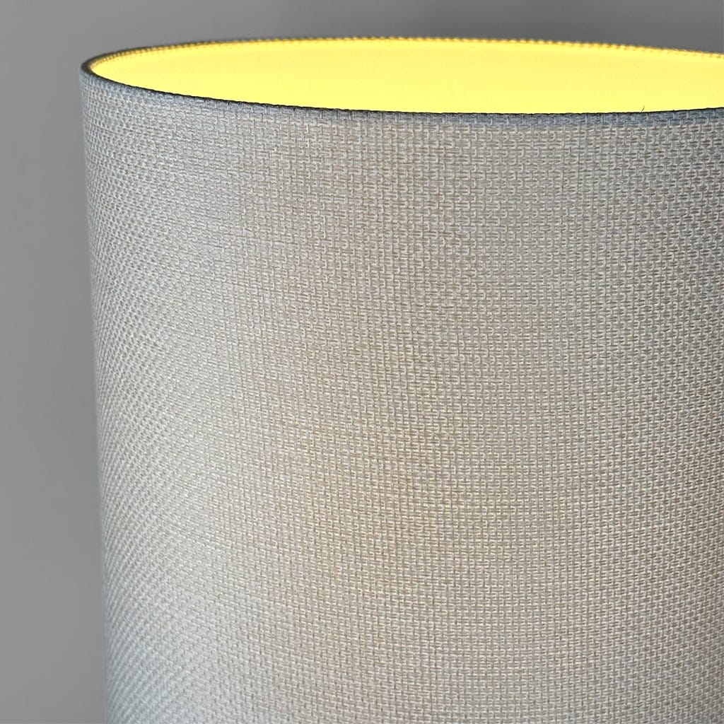 Close-up of a cylindrical lampshade made of woven light beige fabric, showing the textured weave and a warm yellow glow from the interior, set against a neutral gray background.