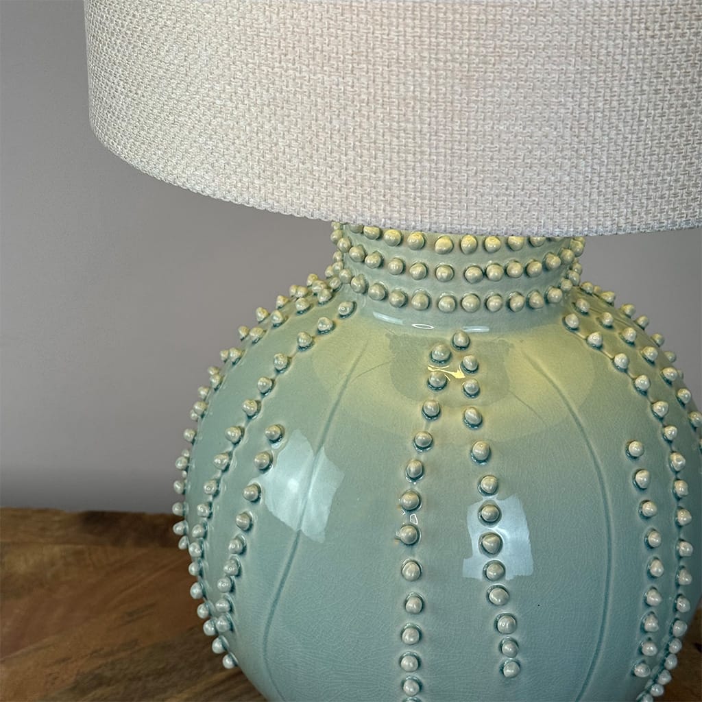 A close-up of a ceramic table lamp with a glossy pale aqua base decorated with raised bead-like details, topped with a textured off-white fabric shade, resting on a wooden surface against a neutral gray wall.