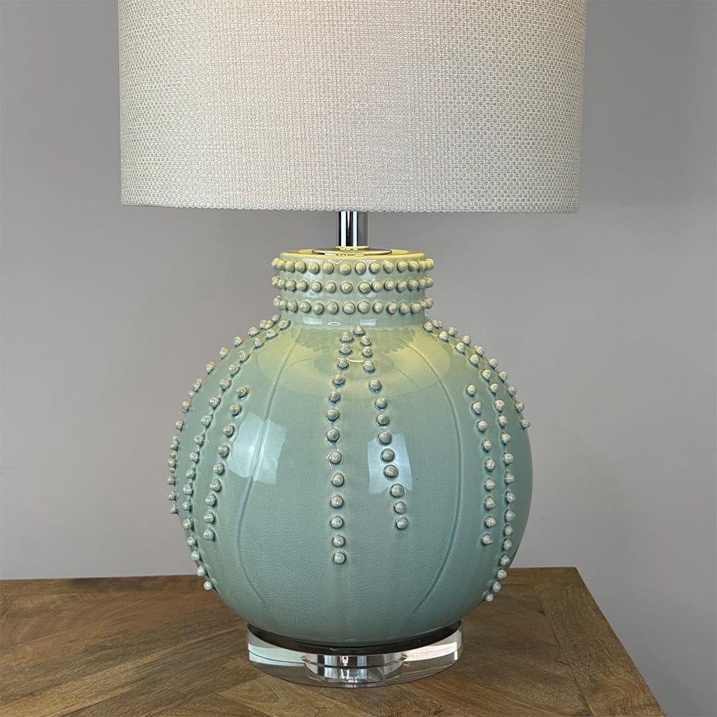 A ceramic table lamp with a rounded, sea urchin-inspired bulb base in a pale sage green glaze, decorated with raised bead-like details, sitting on a wooden table against a gray wall.