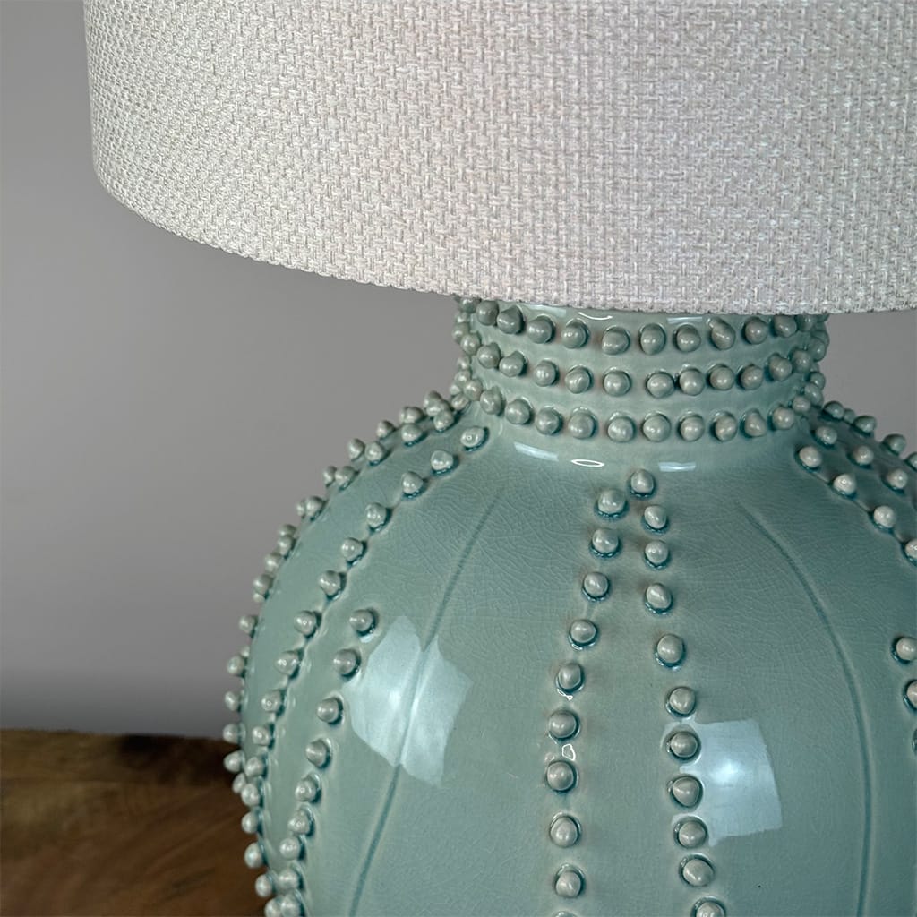 Close-up of a ceramic table lamp base in a pale mint green glaze with raised beaded details, topped with a textured off-white fabric drum shade.