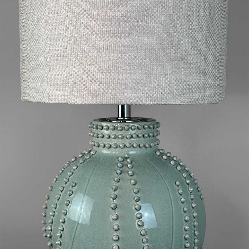 A close-up of a table lamp featuring a glossy pale blue-green ceramic base with vertical rows of raised studs, topped with a textured off-white fabric drum shade.