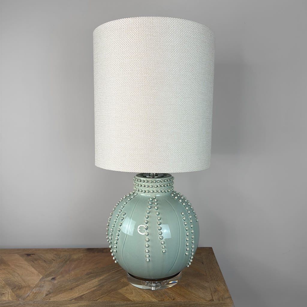 A table lamp with a textured off-white fabric drum shade and a glossy pale aqua ceramic base decorated with raised pearl-like accents, shown resting on a wooden surface against a plain gray wall.