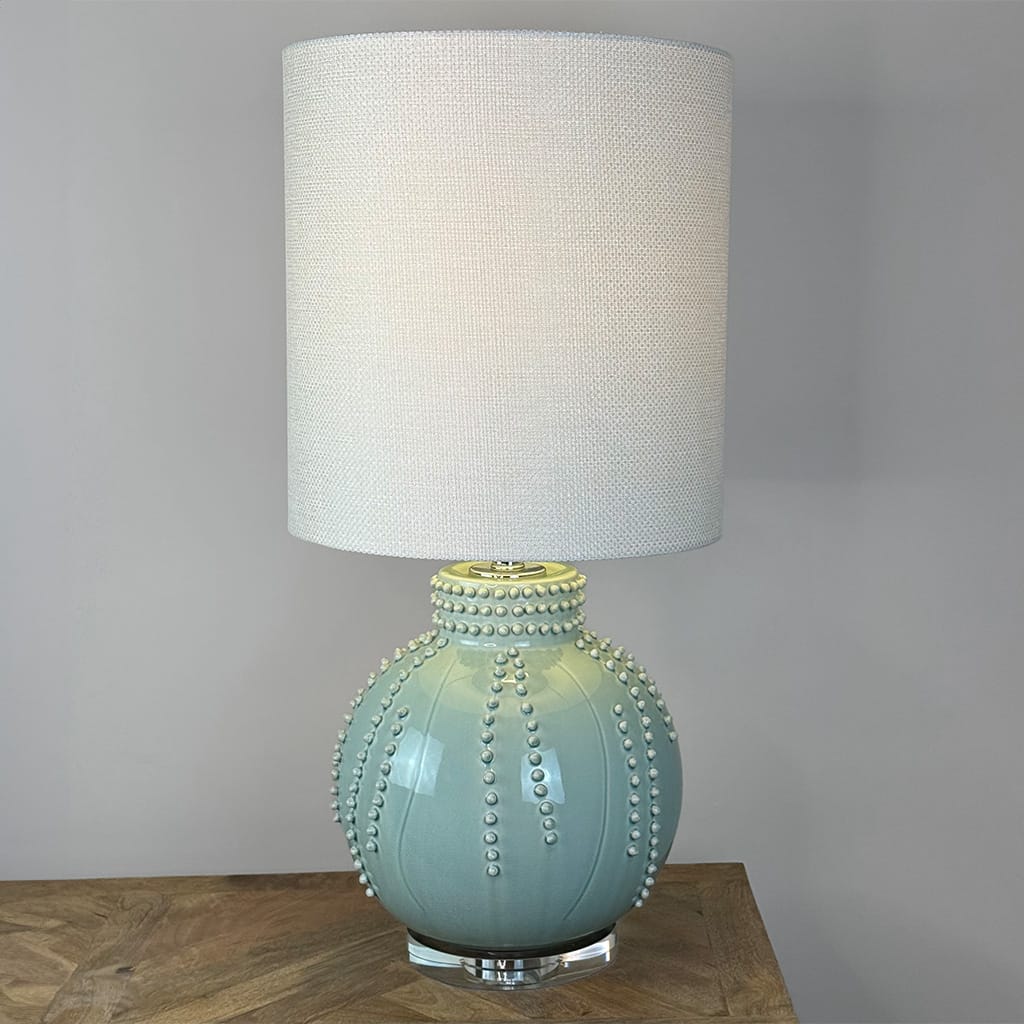 A table lamp with a textured off-white fabric drum shade and a glossy light blue-green ceramic base decorated with raised bead-like details, shown on a wooden surface against a neutral gray wall.
