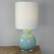 A table lamp with a textured off-white fabric drum shade and a glossy light blue-green ceramic base decorated with raised bead-like details, shown on a wooden surface against a neutral gray wall.
