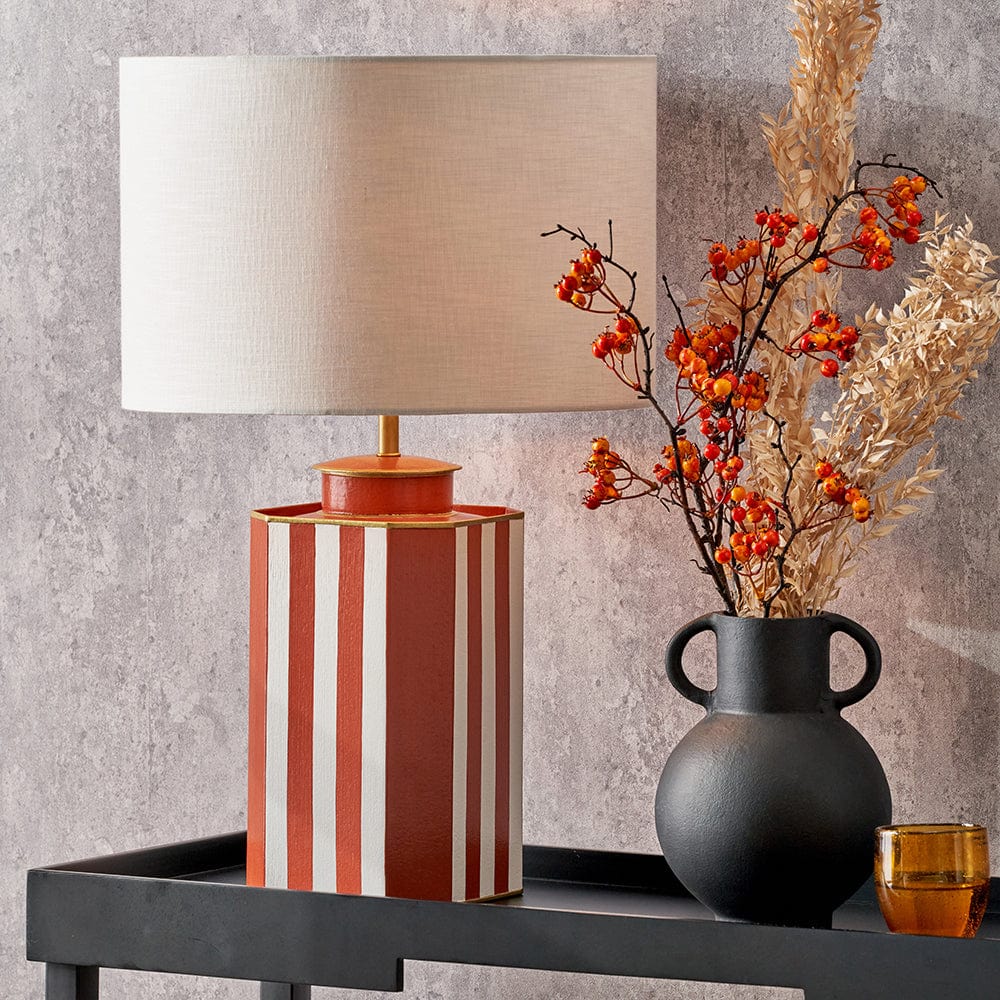 A square metal table lamp base with a hand painted design in white and cinnamon colors, featuring gold accents, positioned on a table with a lamp shade on top.