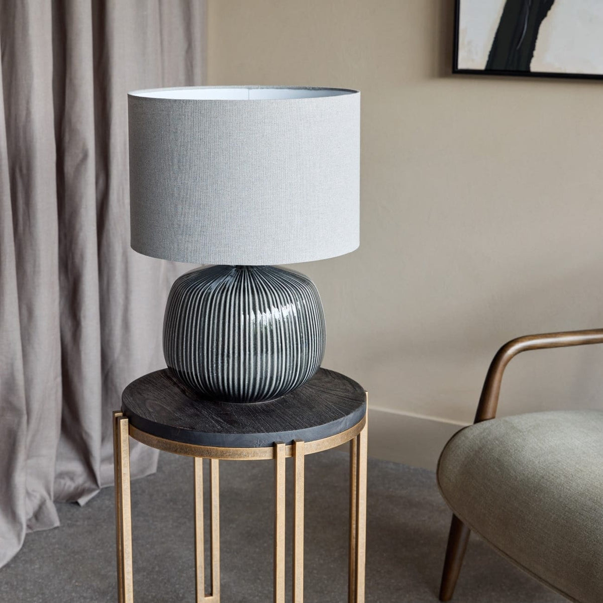 A table lamp with a light gray drum shade and a ribbed dark gray ceramic base, displayed on a small round side table with a dark top and gold metal legs in a styled interior setting.