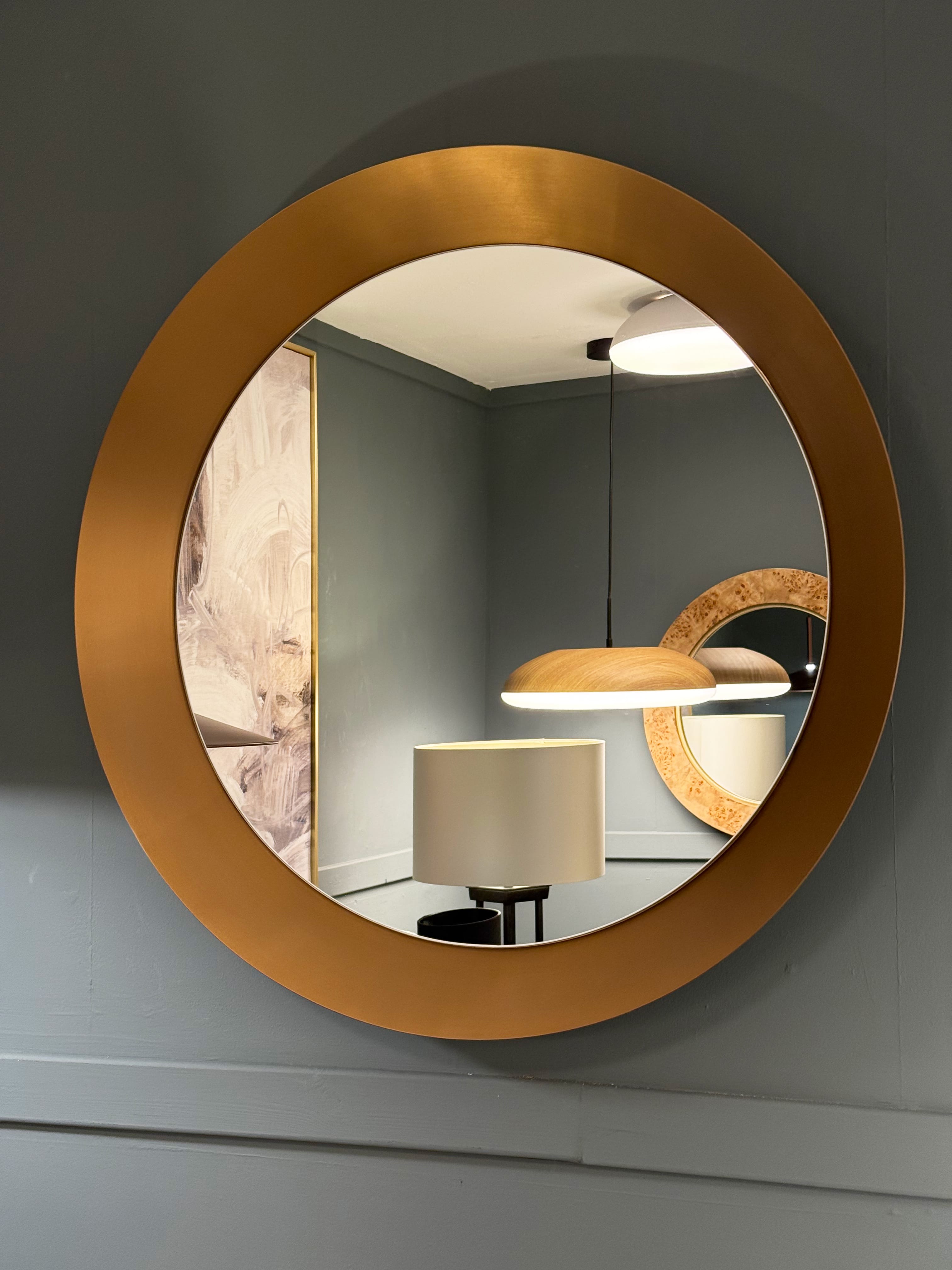 A large round wall mirror with a wide brushed gold frame, mounted on a dark gray wall. The mirror reflects a modern room interior with a pendant light and a lamp.