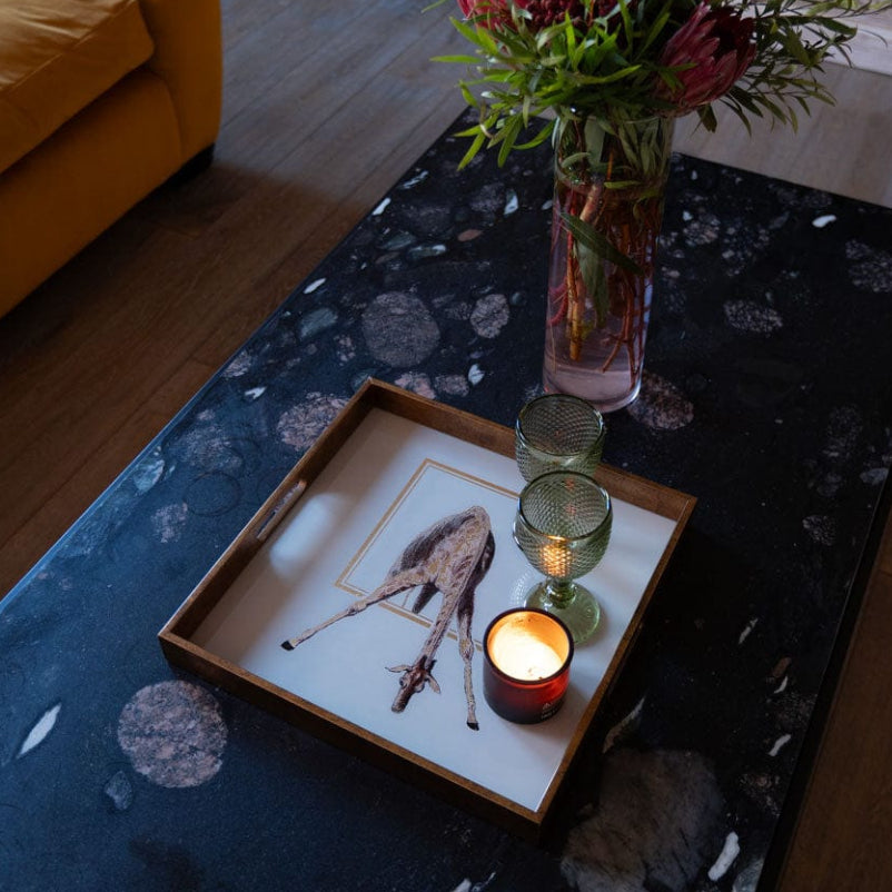 A square giraffe tray with gold tones, placed on a table with a floral pattern, serving as a platter for a candle and a flower vase.
