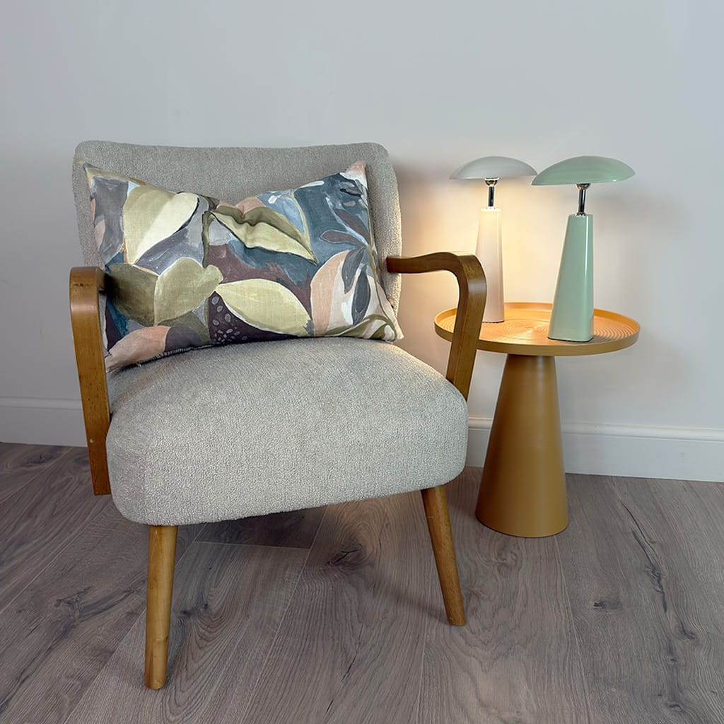 Gray armchair with a patterned pillow next to a small round table with two lamps on a wooden floor.