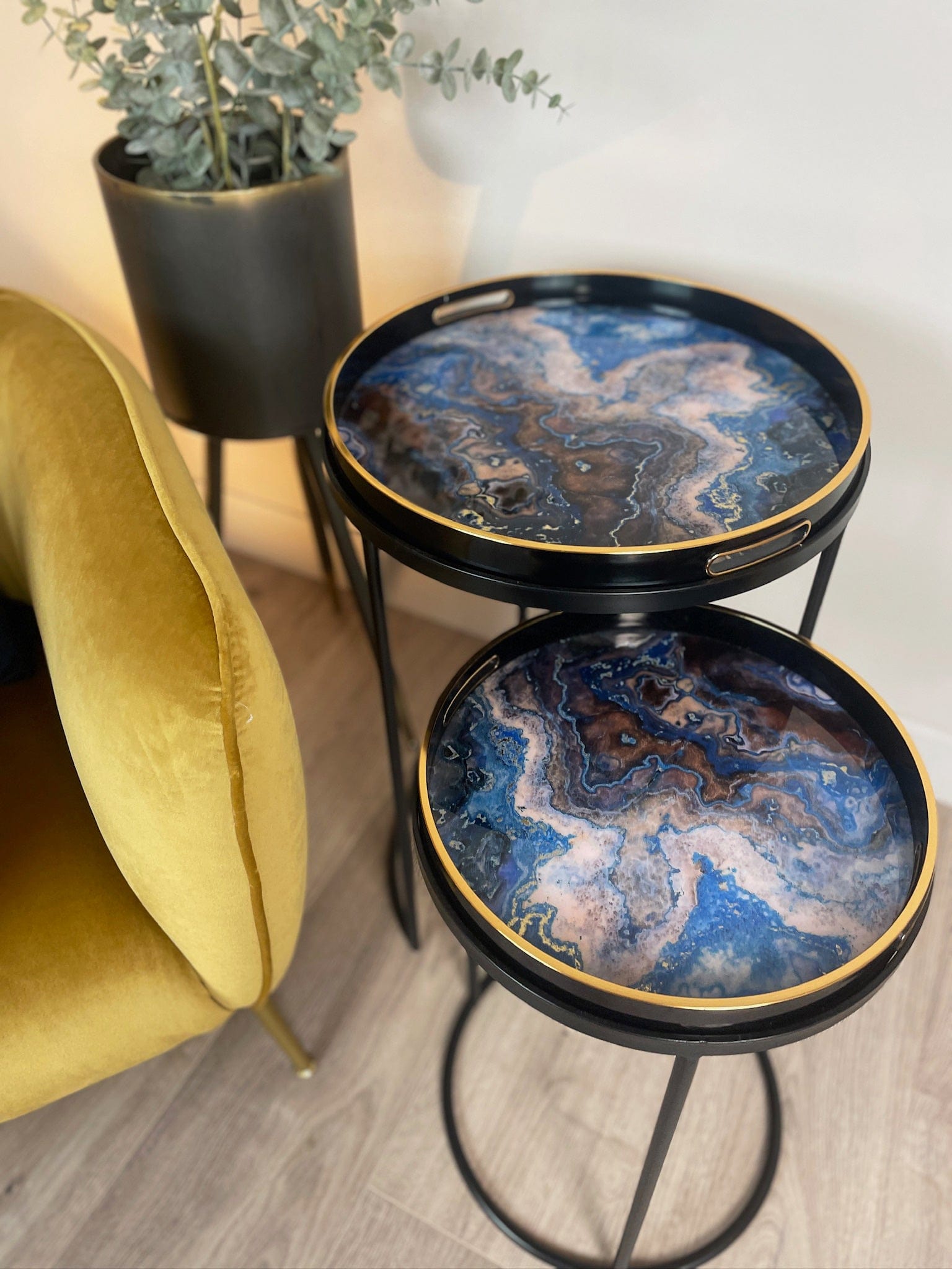 Two-tiered decorative table with marble-like design in a room setting with a yellow chair and plant.