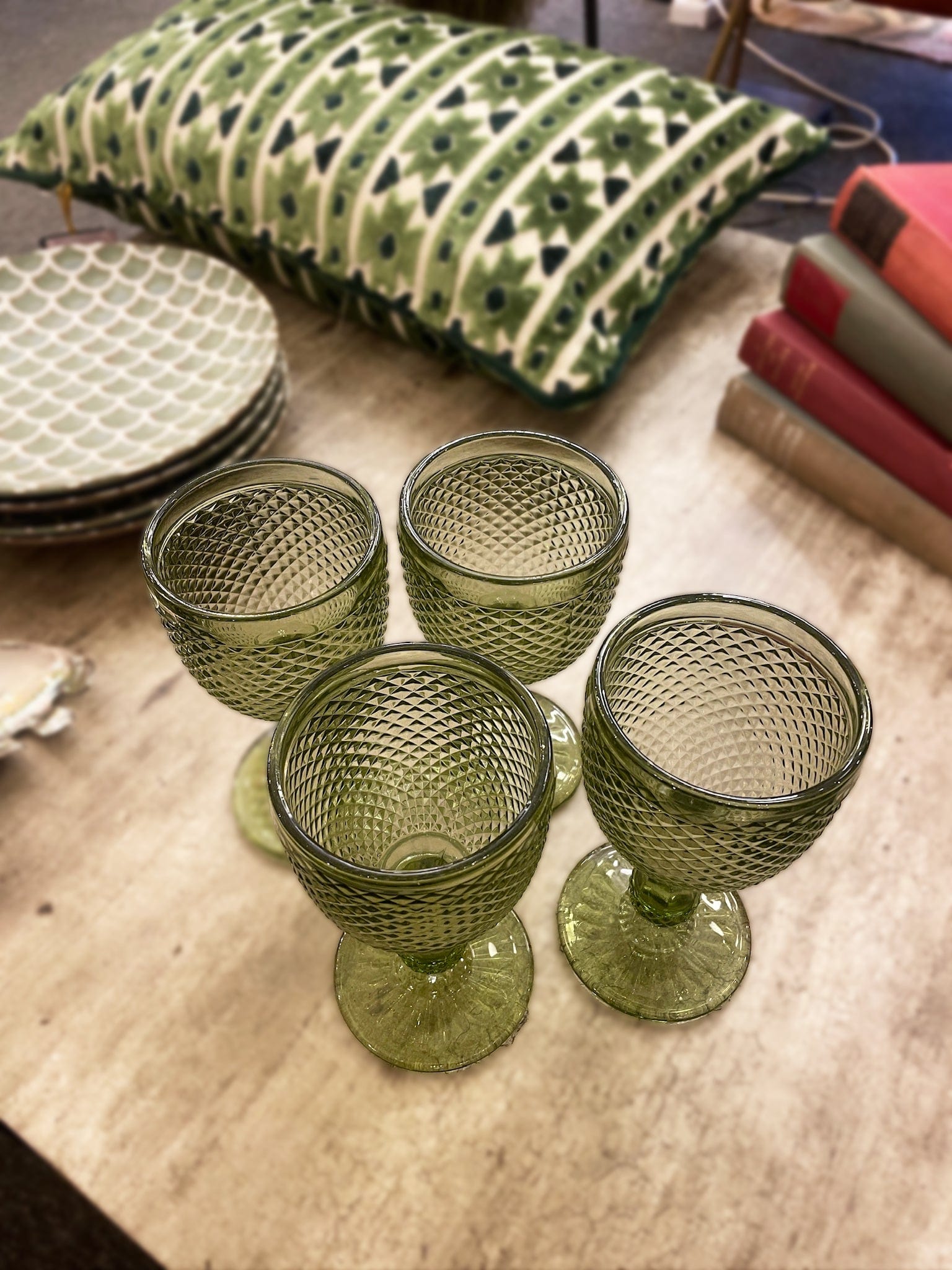 Set of four green textured glass goblets on a wooden surface with a patterned pillow and books in the background.