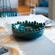 A teal ceramic bowl with a glossy finish and decorative balls around the rim, placed on a dining table.