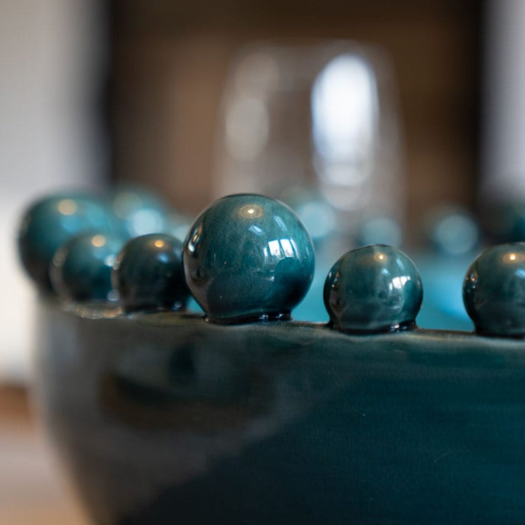 A teal ceramic bowl with a glossy finish and decorative balls around the rim with a blurred background