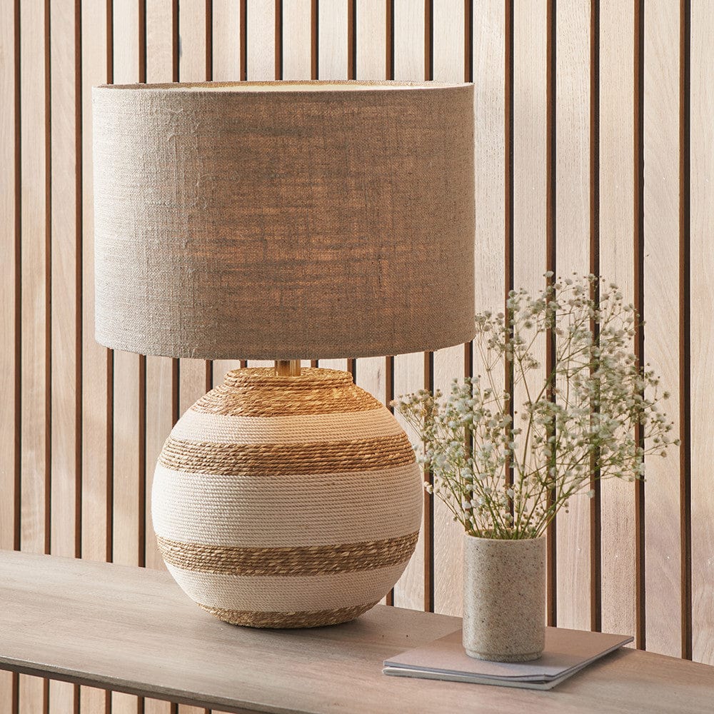 A round table lamp with a beige and natural sea grass base, placed on a wooden surface with a vase of flowers beside it.