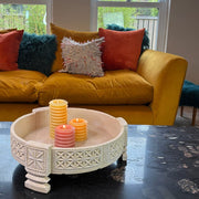 Decorative footed bowl with candles on a dark surface in front of a yellow couch with colorful pillows.