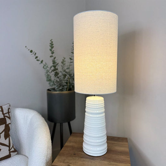 White textured table lamp on a wooden surface with a plant in the background