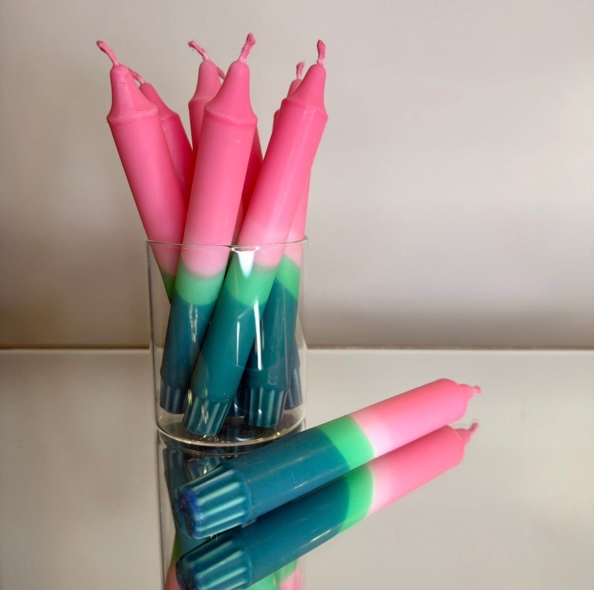 Pink and green candles in a glass holder on a reflective surface with a neutral background
