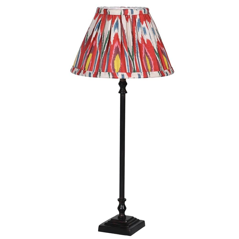 A black metal stem candlestick lamp with a hand made pleated small empire shaped lampshade featuring an Ikat pattern in tones of red, pink, violet, blue, and yellow.