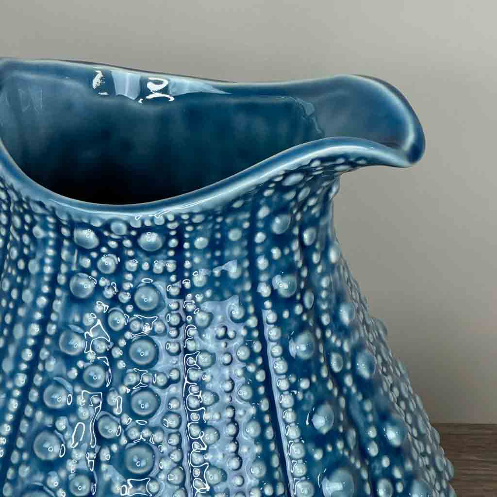The spout of a large blue ceramic jug with a textured surface, resembling the form of a sea urchin, displayed against a grey wall