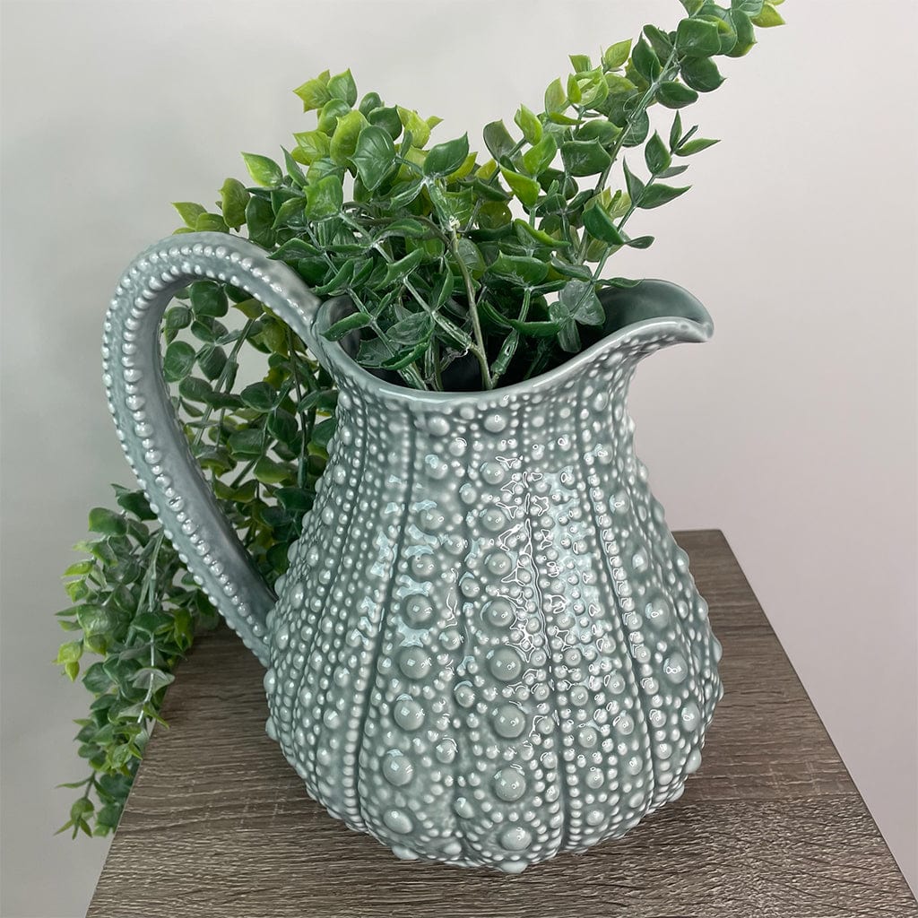A large grey ceramic jug with a textured surface, resembling the shell of a sea urchin, displayed on a wooden surface with some faux greenery inside.