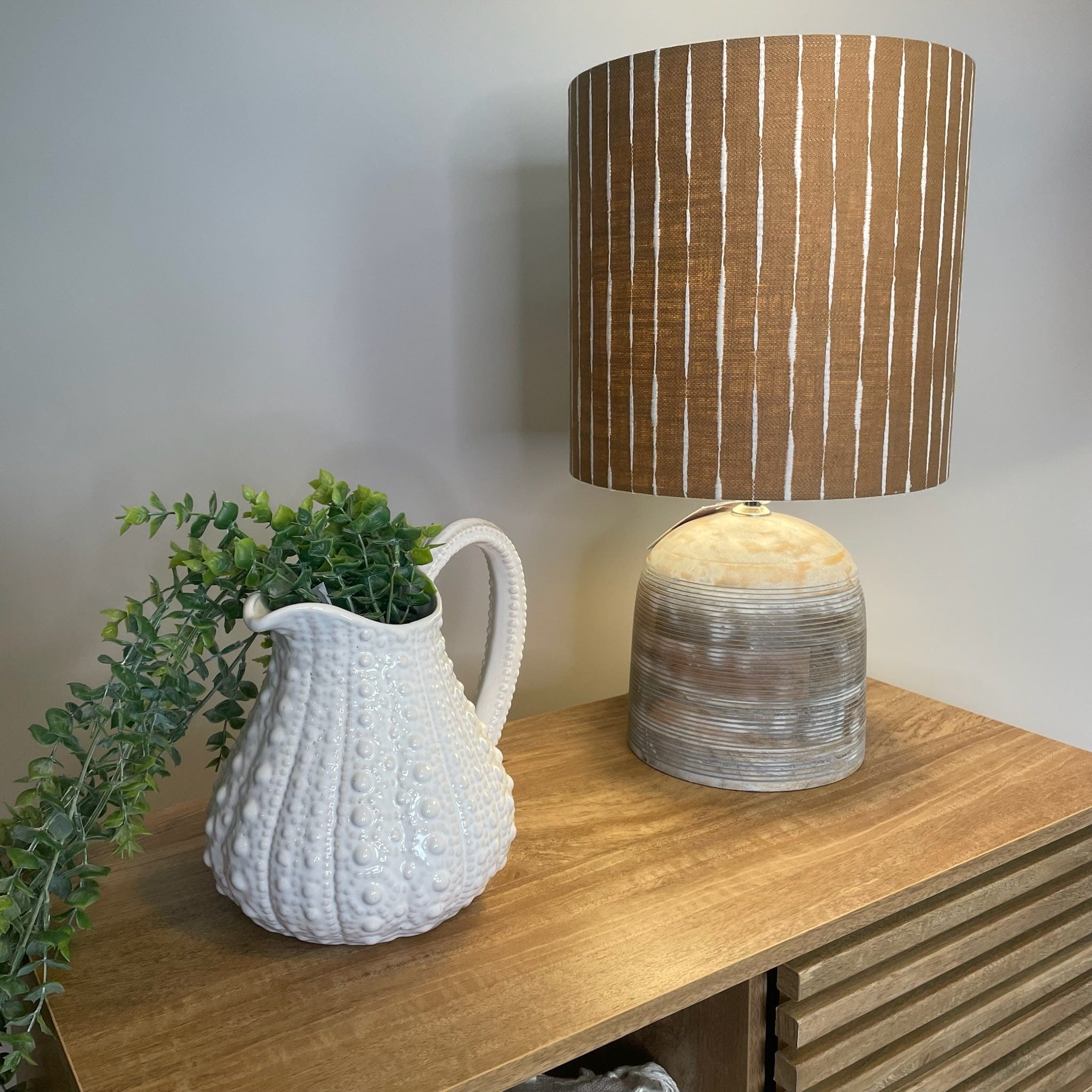 A large white ceramic jug with a textured surface, resembling the shell of a sea urchin, displayed with faux greenery inside next to a wooden table lamp with a tall striped shade in a tan and white tone.