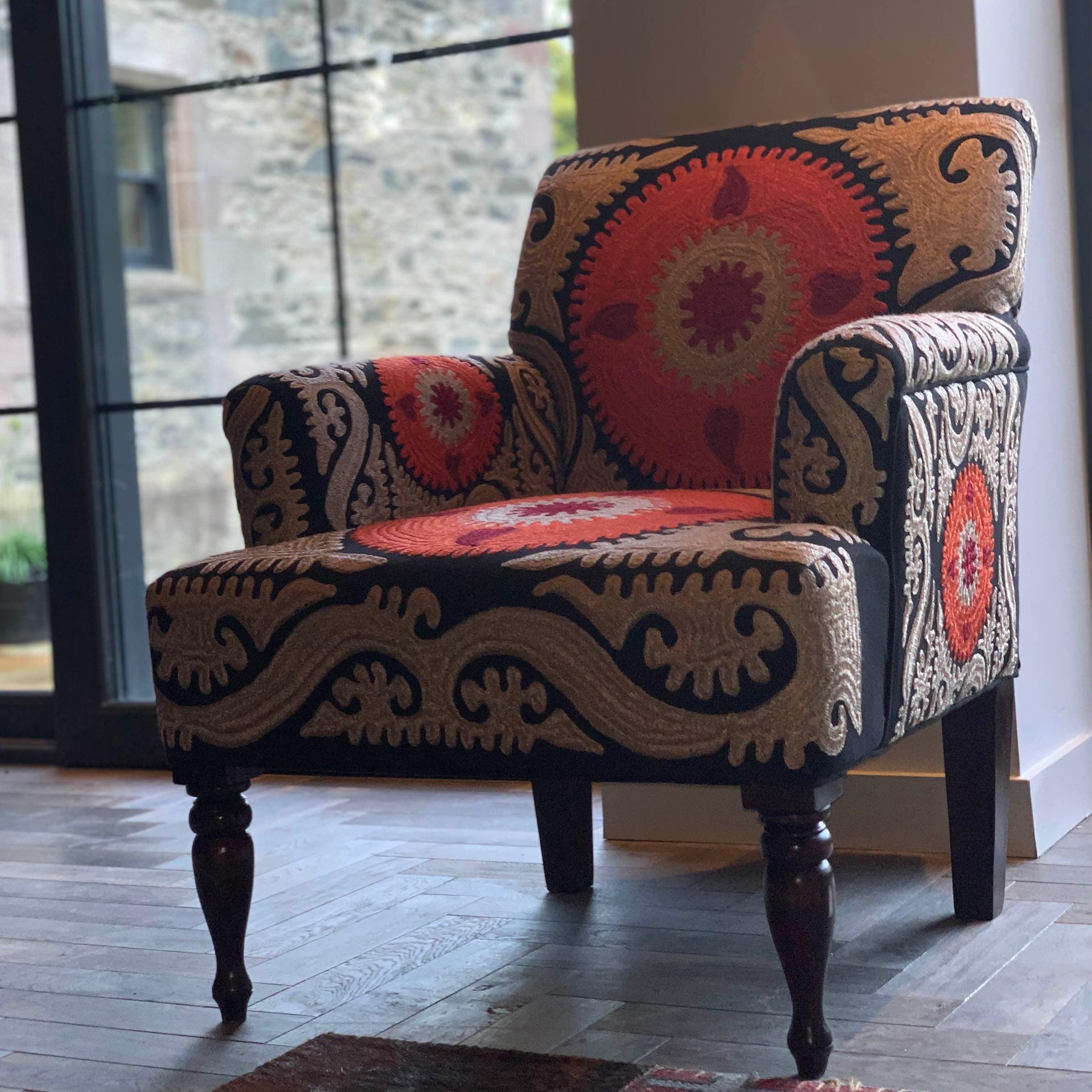 An ornate armchair with multicolored floral and geometric embroidery on the upholstery, standing on a hardwood floor near a large window.