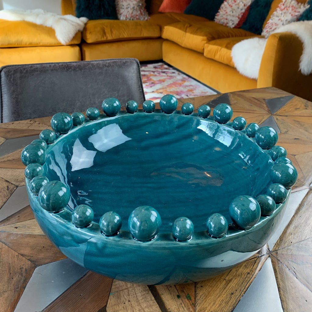 A teal ceramic bowl with a bobbled edge containing spherical decorations, placed on a wooden surface with a cozy room background featuring a yellow sofa and a gray chair.