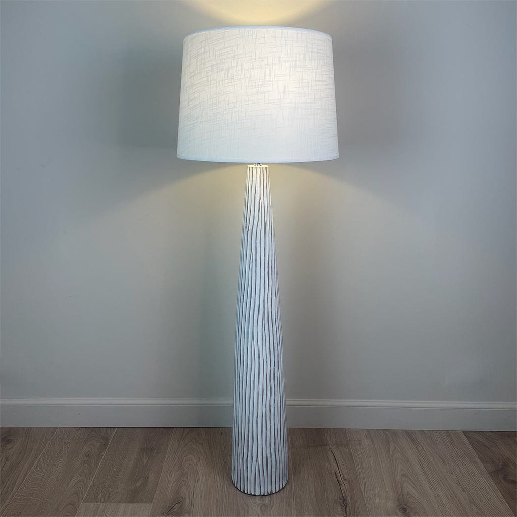 A white wood effect tall and slim floor lamp with a large white linen shade and the light is on