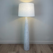 A white wood effect tall and slim floor lamp with a large white linen shade and the light is on