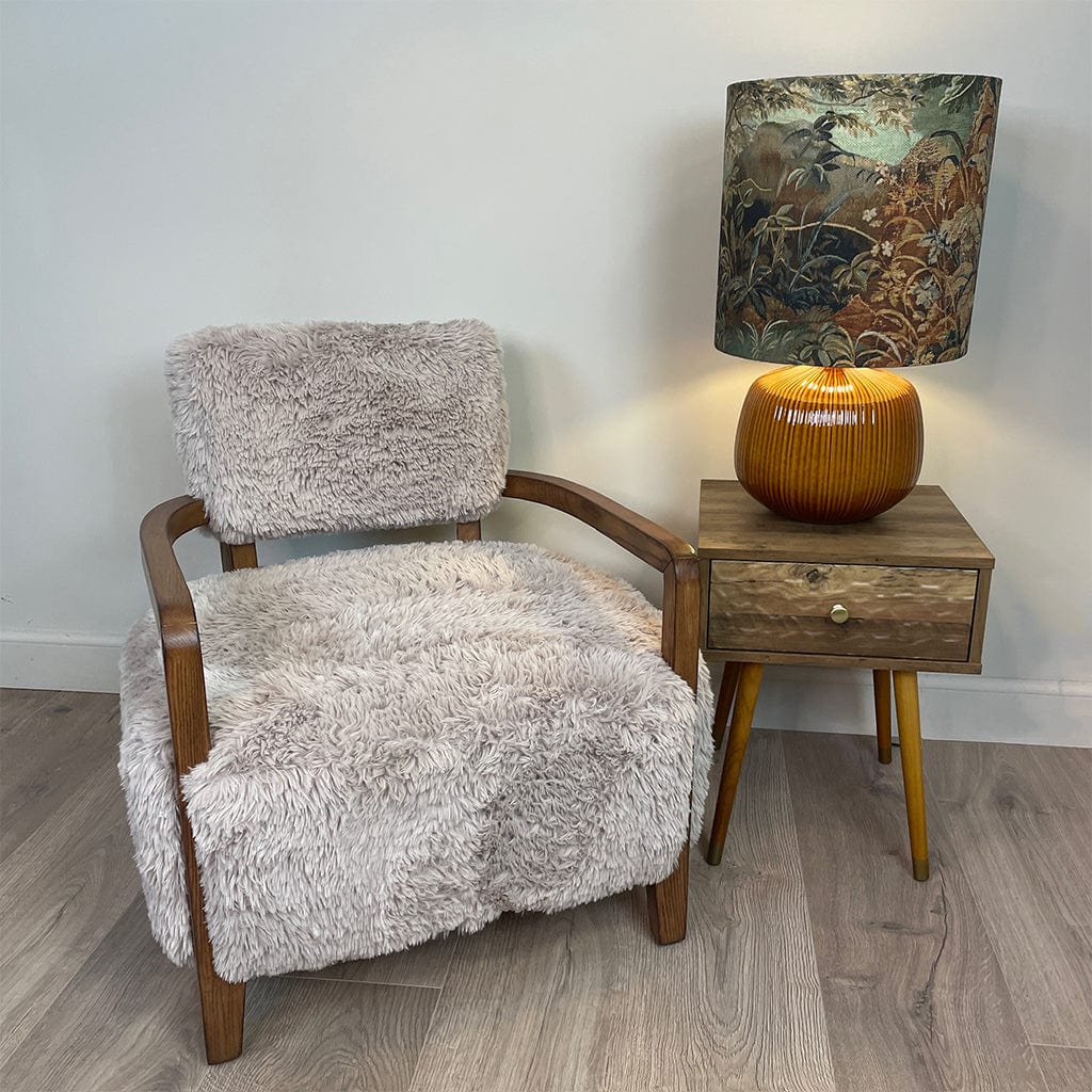 A caramel-colored faux fur armchair with dark ash wood arms, displayed in a cozy indoor setting next to a wooden side table with an ochre lamp and a tall lampshade with a forest scene. 